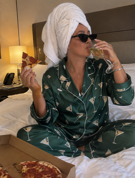 A woman on a hotel bed in satin pyjamas with champagne and pizza