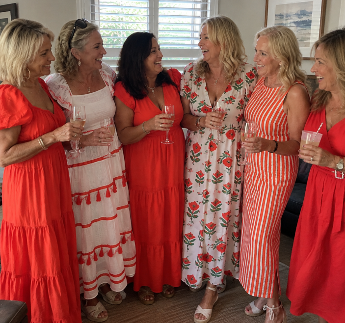 Sandra: Six women in coordinated red and white outfits standing together indoors holding champagne and cocktails, laughing with each other rather than at the camera
