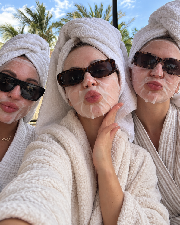 Three women in robes and sheet masks outdoors under palm trees