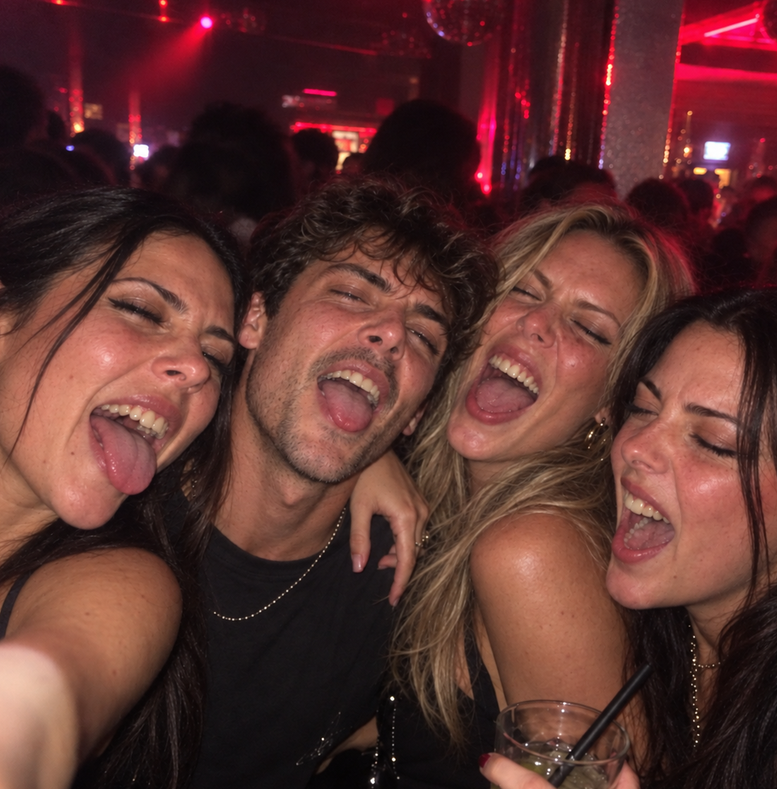 Brooke: Four friends packed together taking a selfie on a nightclub dance floor under red lighting, three women and one man, all pulling faces with their tongues out, one holding a drink