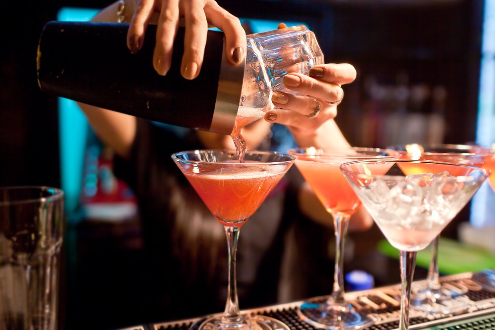 A shaker pouring a pink cocktail into a row of martini glasses