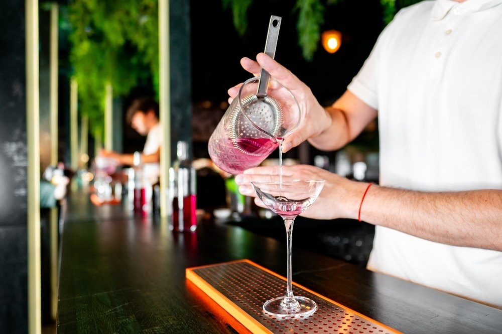A bartender straining a pink cocktail into a coupe glass at a lit bar