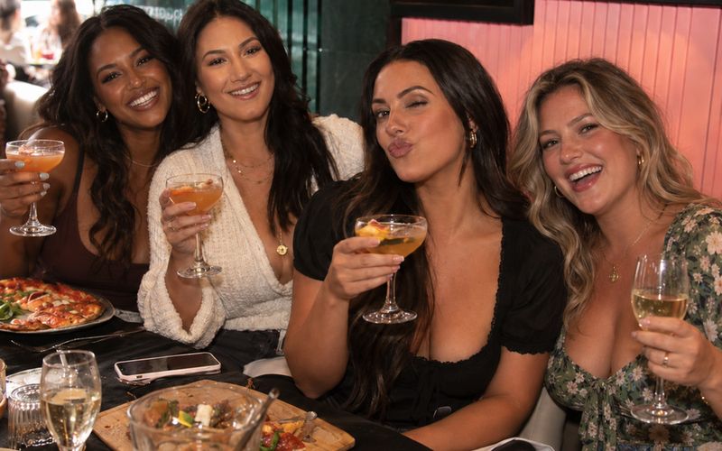 Four friends raising cocktails over food at a busy table on a Melbourne girls weekend