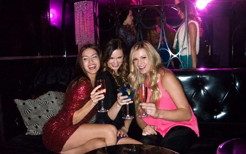 Three friends laughing and raising drinks in a nightclub booth on a Sydney girls weekend