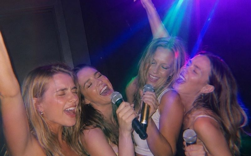 Four friends belting out a song together into microphones in a private karaoke suite on a Sydney girls weekend