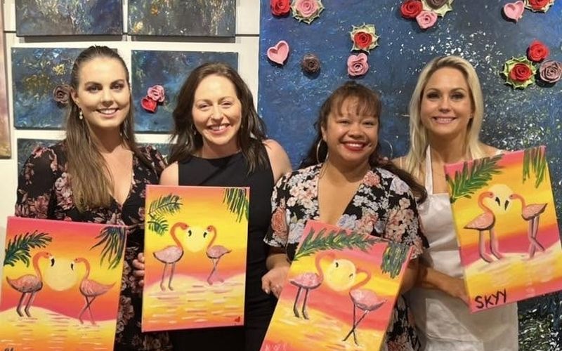 Four friends proudly holding up their flamingo sunset paintings on a Brisbane girls weekend