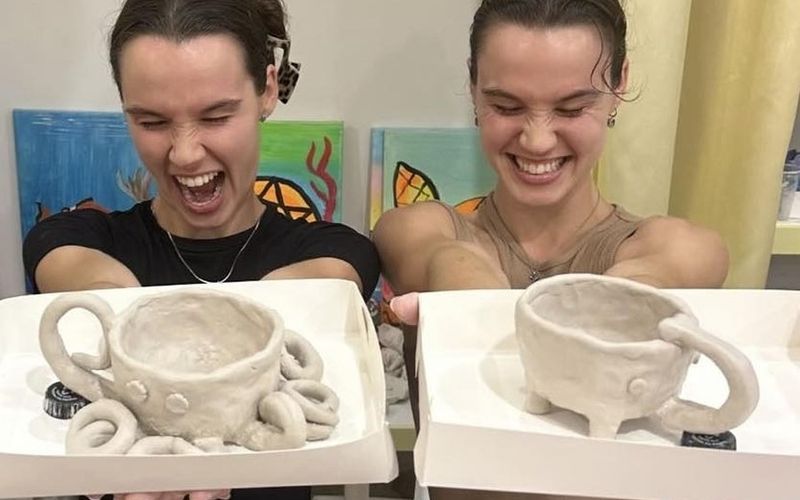 Two friends laughing as they hold up their handmade clay mugs on a Brisbane girls weekend