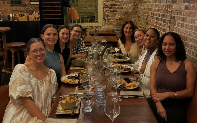The girls smiling along a long timber table in a heritage brick pub on a Brisbane girls weekend