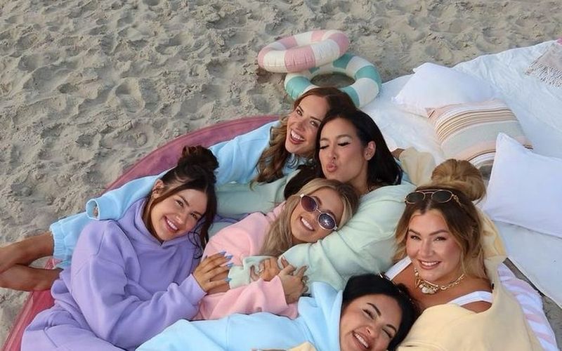 Six friends in pastel hoodies piled onto a beach float on the sand, laughing on a Brisbane girls trip
