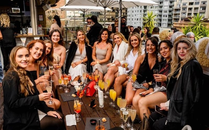 A big group of friends toasting drinks around a rooftop table on a Brisbane girls weekend