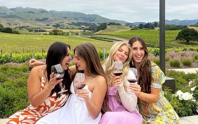 Four friends laughing with red wine on a terrace wall overlooking the vines on a Brisbane girls trip