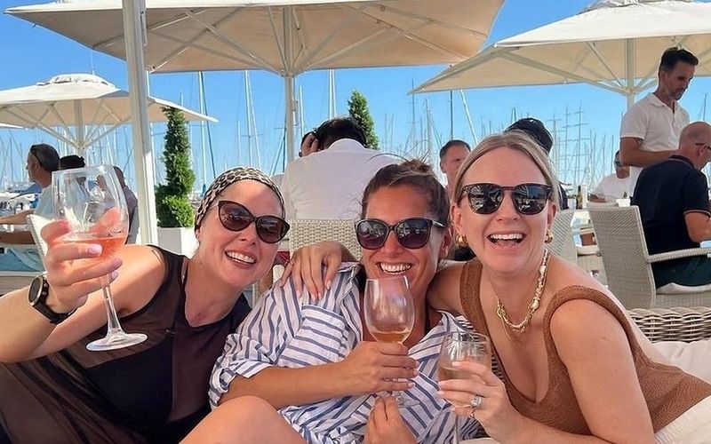 Three friends in sunglasses laughing with drinks under the umbrellas at a beach club on a Gold Coast girls weekend