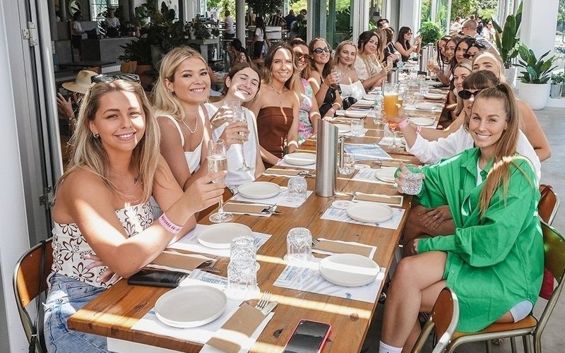A long table of friends smiling over mimosas at a bright lunch on a Gold Coast girls weekend