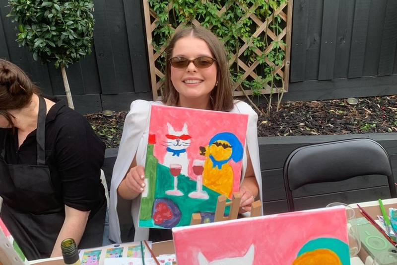 A painter in sunglasses holding up her painting of a cat and a dog in sunglasses
