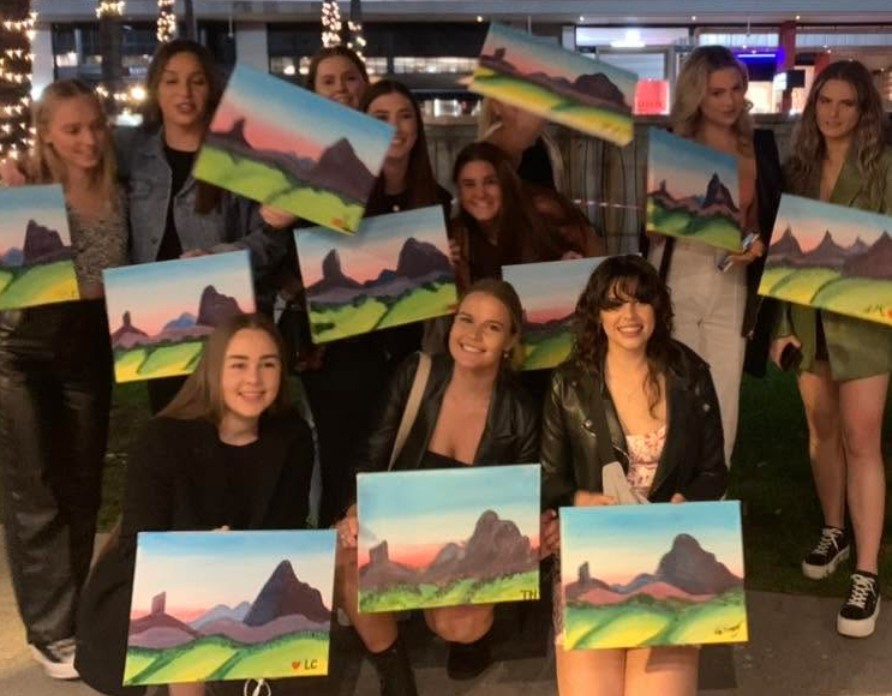 A big group holding up their mountain paintings outside under fairy lights at night