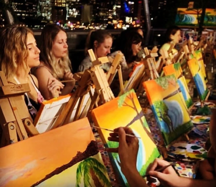 A row of painters at easels painting bright sunsets, the city lights glowing behind them at night