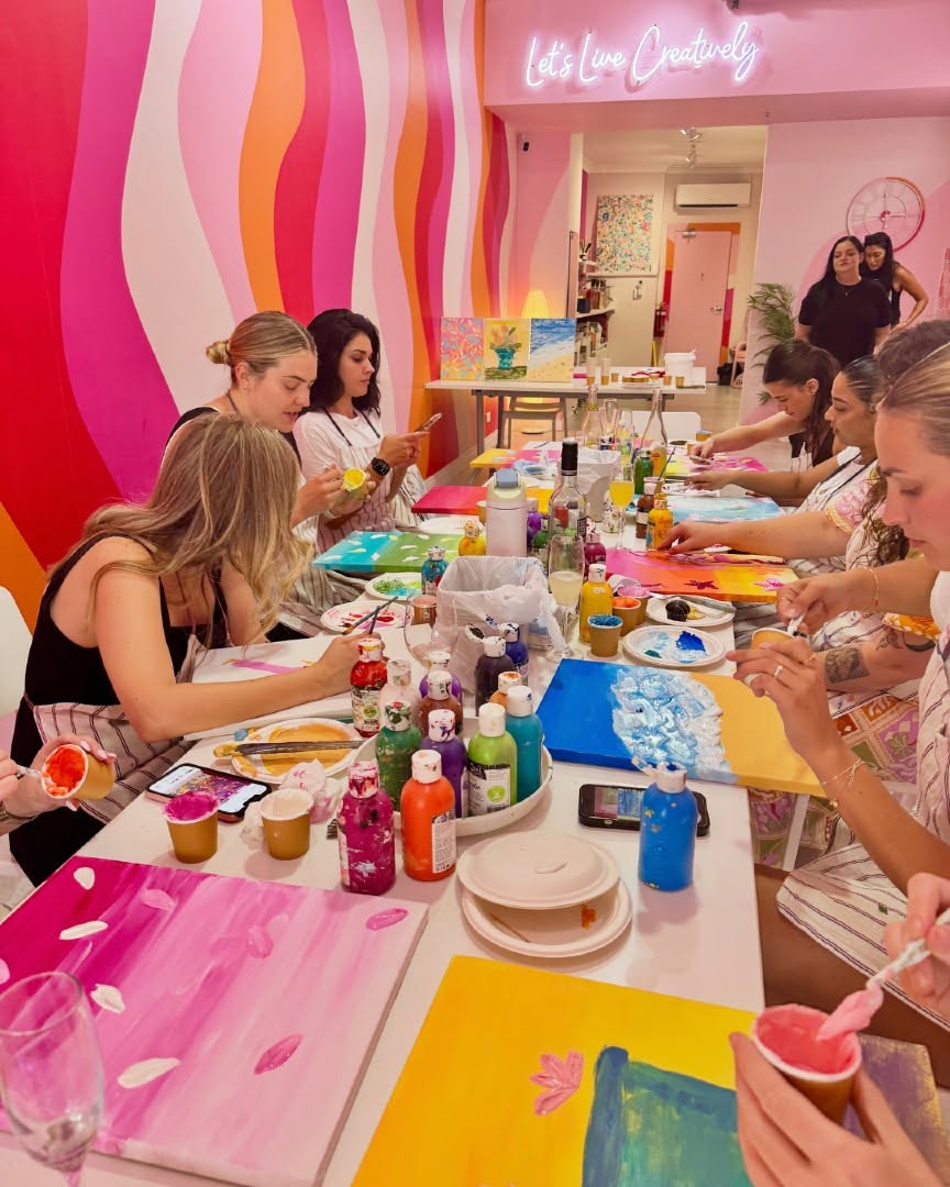 Friends painting bright canvases along a long table in a studio with a pink and orange mural