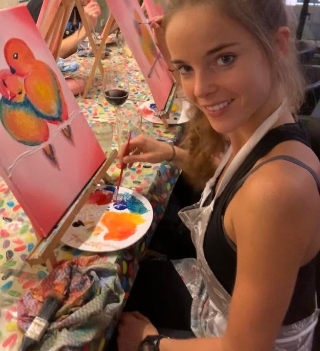 A painter in an apron smiling beside her painting of two lovebirds, a glass of red at her elbow