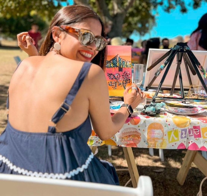 A painter in sunglasses laughing over her shoulder at an outdoor painting table