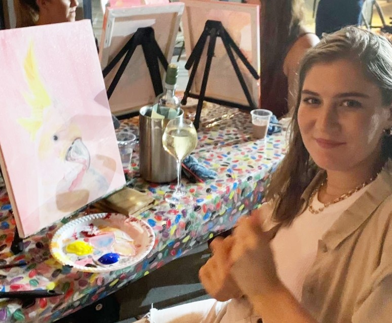 A painter beside her soft pink cockatoo painting, a glass of white in hand
