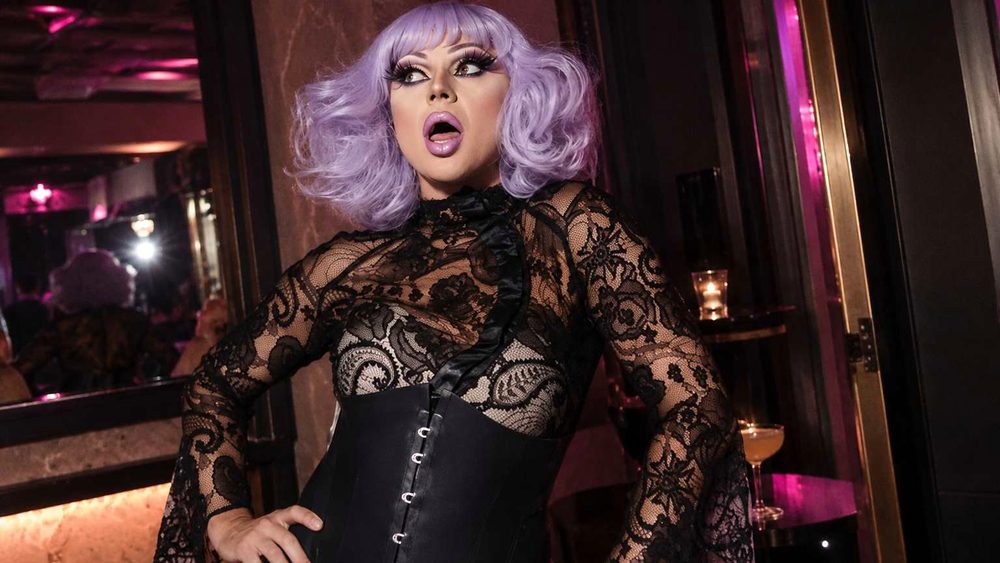 A drag performer in a lilac wig and black lace striking a surprised pose in a pink-lit bar