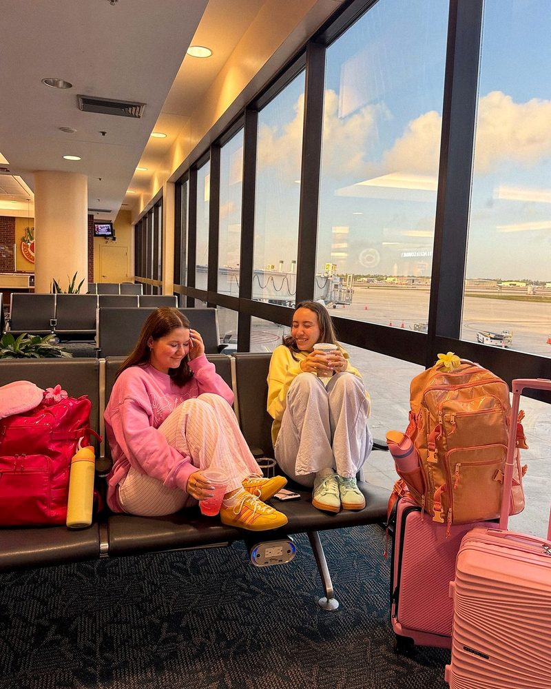 Two friends with coffees and pink luggage at an airport gate at sunrise