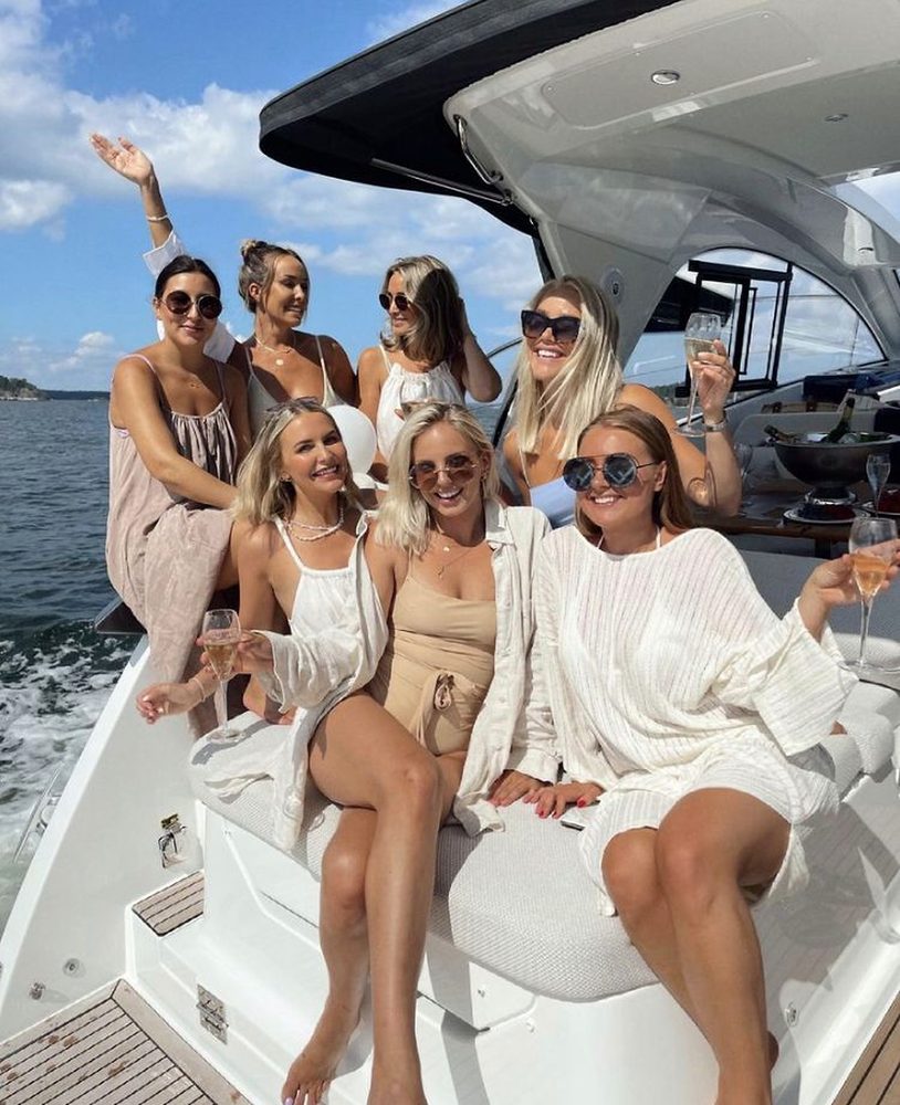 Seven friends in summer dresses laughing with champagne on the back of a boat