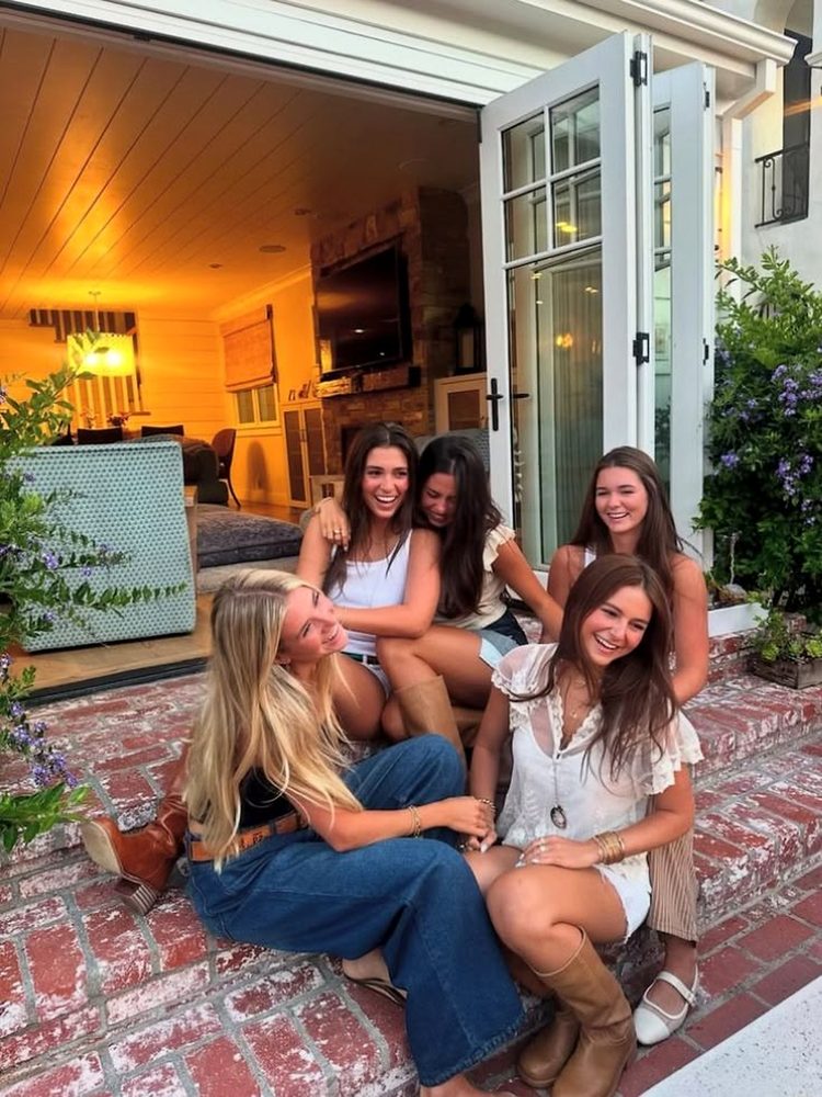 Five friends laughing together on red brick steps outside open doors into a warm lit living room