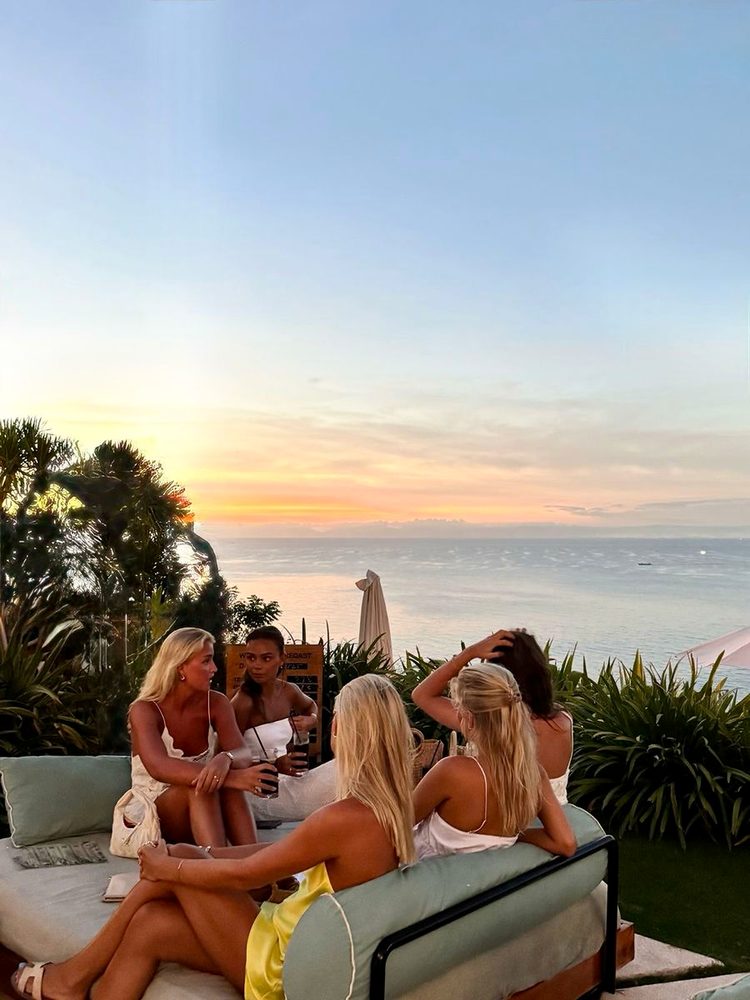 Six friends chatting with drinks on a clifftop terrace above an ocean sunset