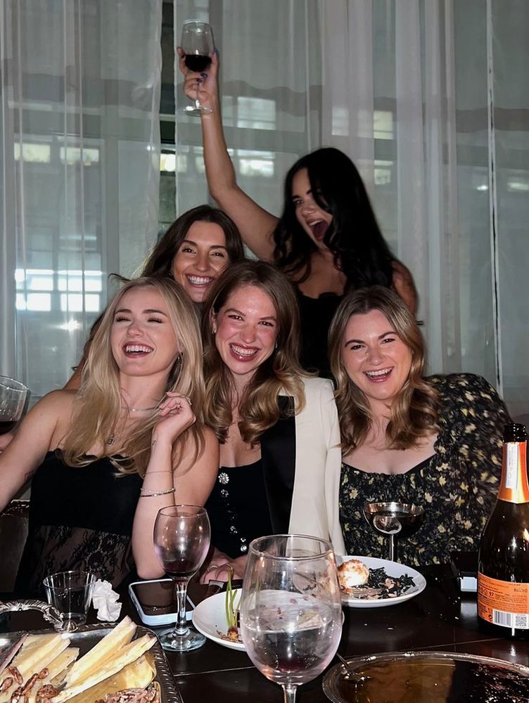 Five friends laughing at a dinner table with red wine and a cheese board, one raising her glass