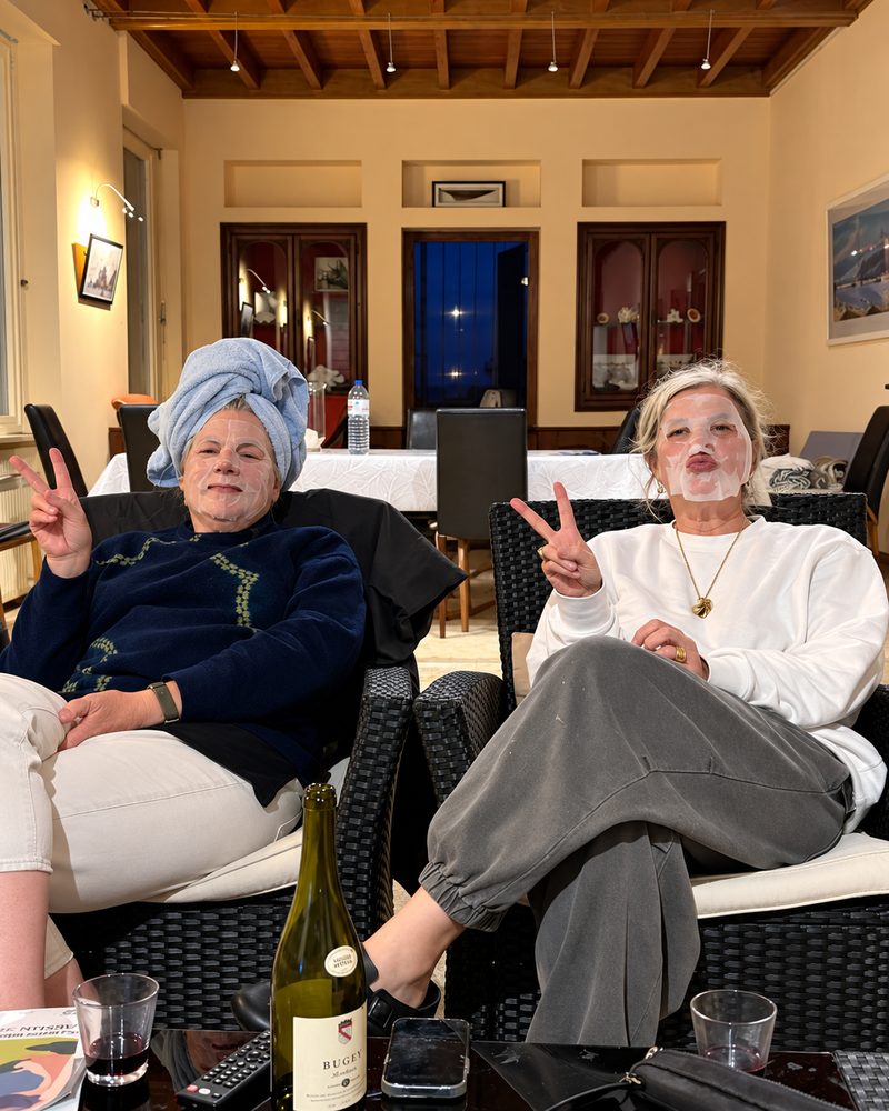 Two friends in their sixties in sheet face masks making peace signs over a glass of wine
