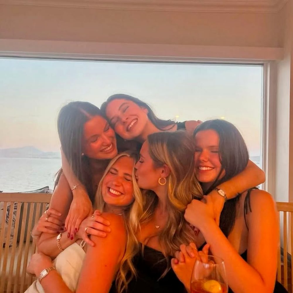 Friends in a group hug by a sunset window with spritzes in hand