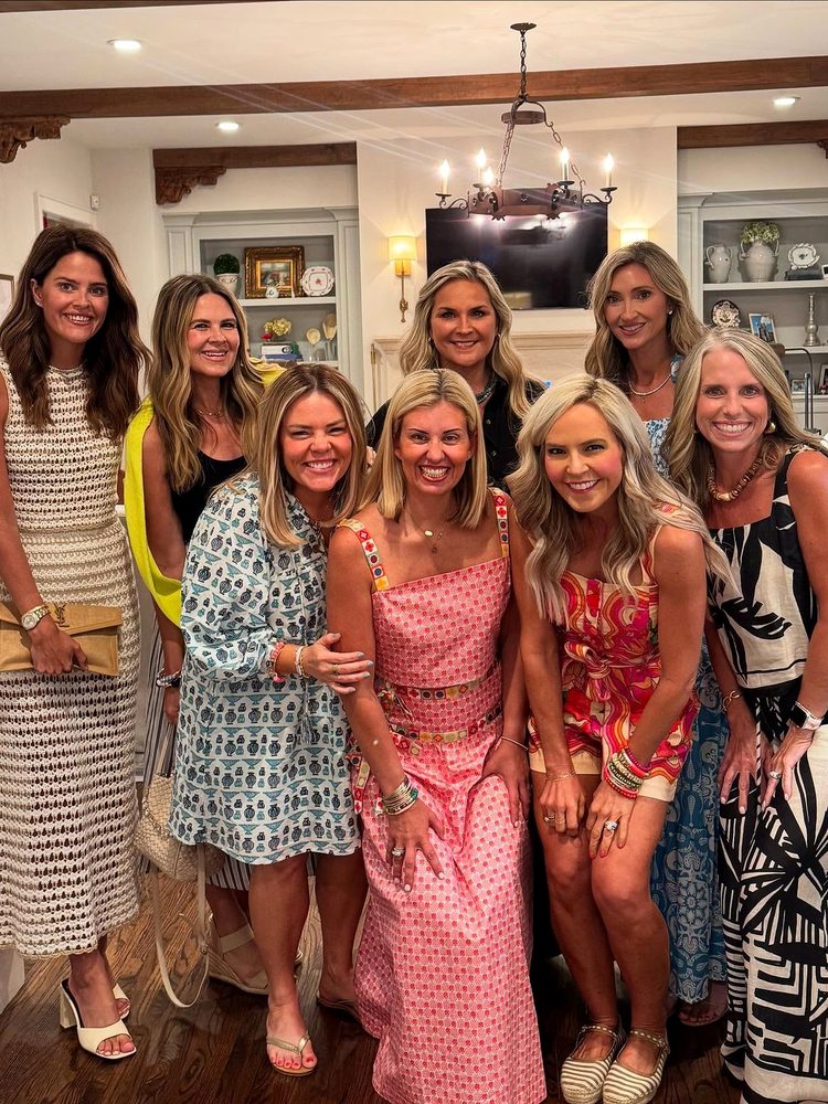 Eight women in their forties and fifties in colourful dresses posing close together in a living room