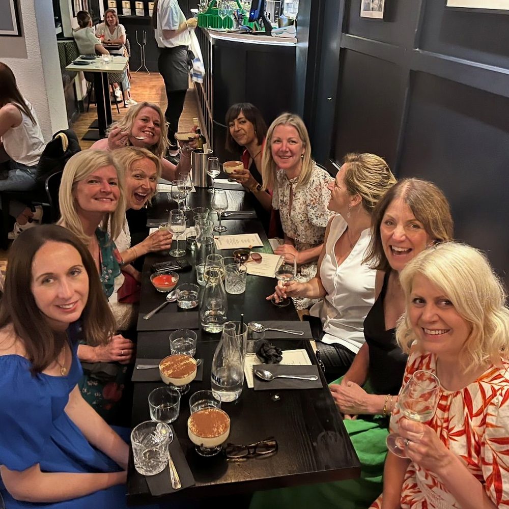 Nine women smiling down a long restaurant table over dessert and wine