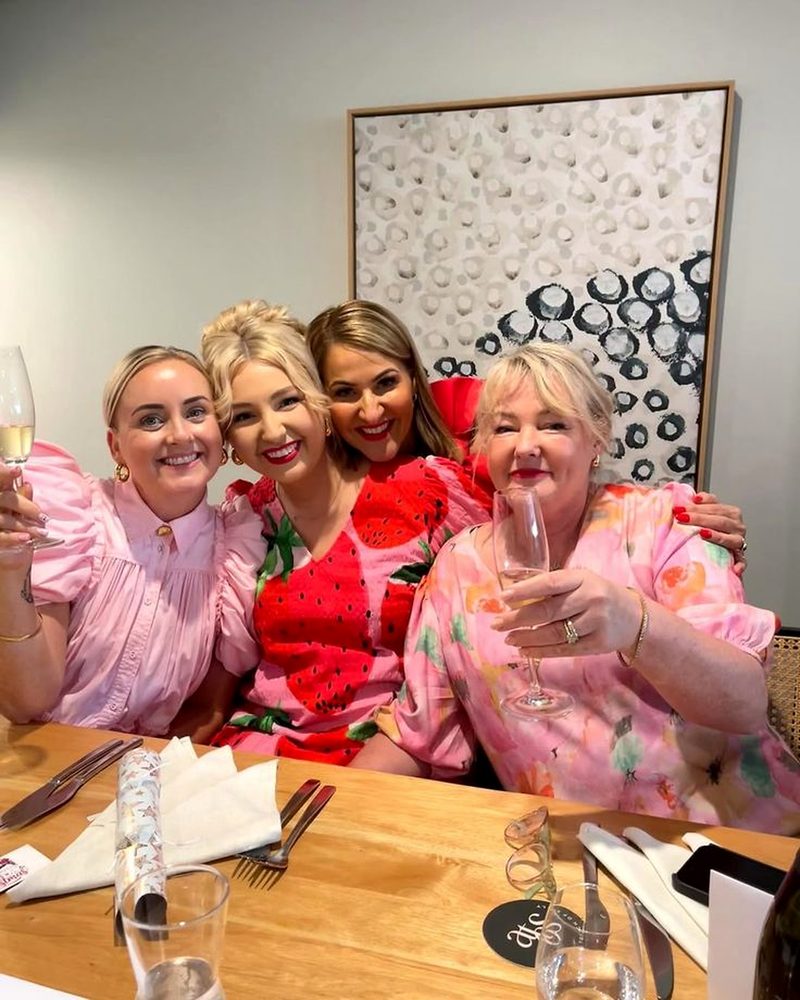 Four women in bright printed dresses smiling together over a long lunch with sparkling wine