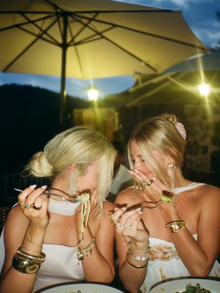Two friends laughing into their hands over pasta at an outdoor dinner