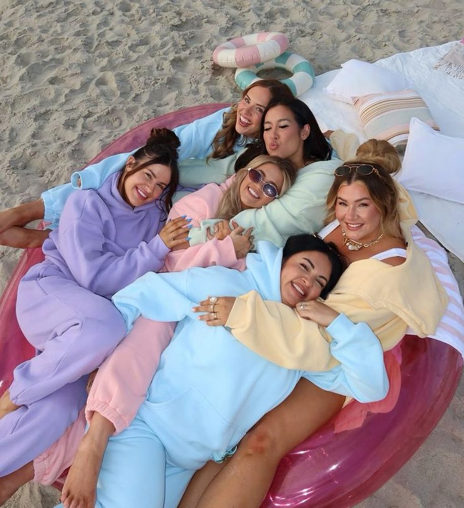 Seven friends in matching pastel tracksuits piled together on a pink float on the sand