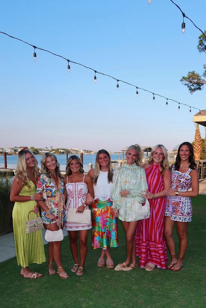 Seven friends in bright resort dresses arm in arm on a waterfront lawn under festoon lights