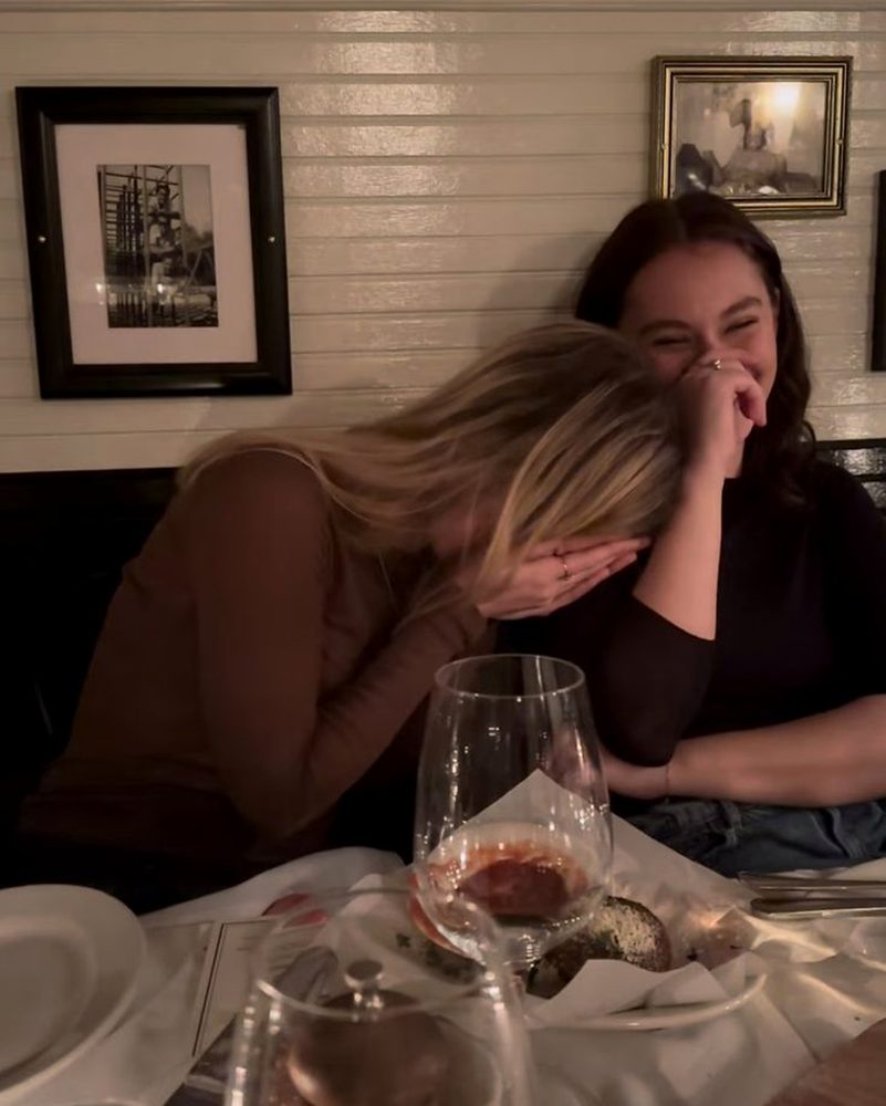 Two friends doubled over laughing in a restaurant booth with red wine