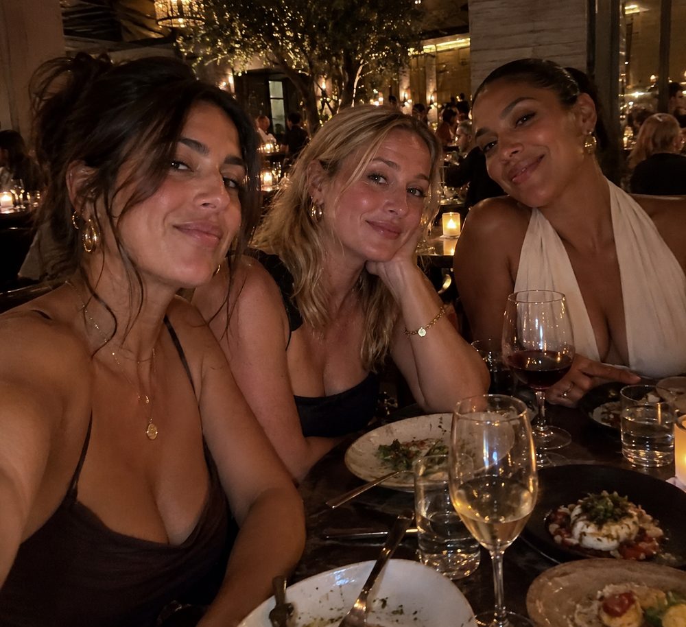 Three friends smiling for a selfie at a candlelit restaurant table with wine and shared plates