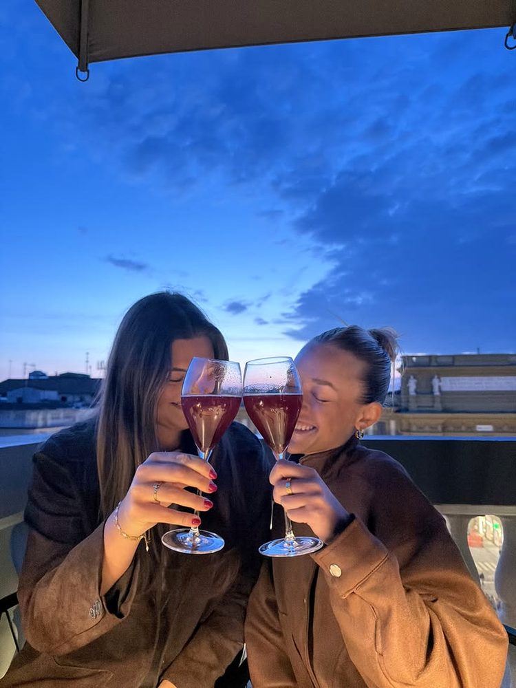 Two friends cheek to cheek clinking red sparkling wine on a city rooftop at dusk
