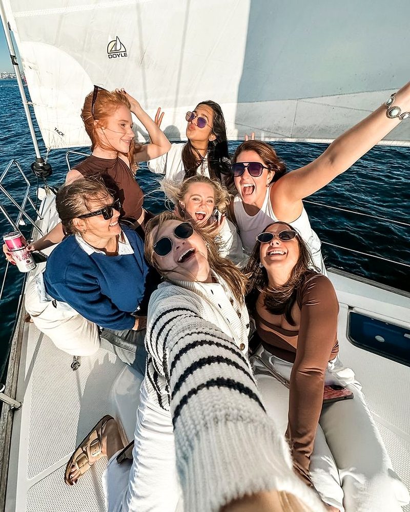 Eight friends laughing in a wide group selfie on the deck of a sailing yacht