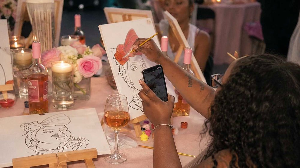A woman painting a line portrait on canvas and photographing it, with rose and flowers on the table