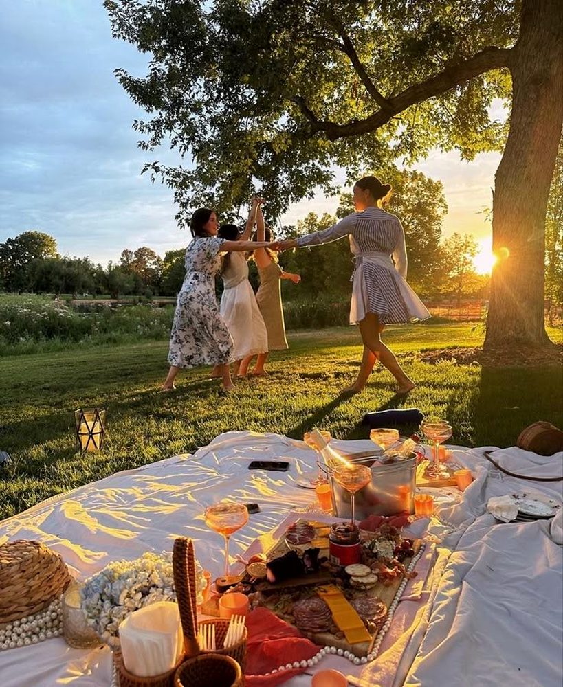 Four friends dancing hand in hand under a big tree at golden hour beside a picnic