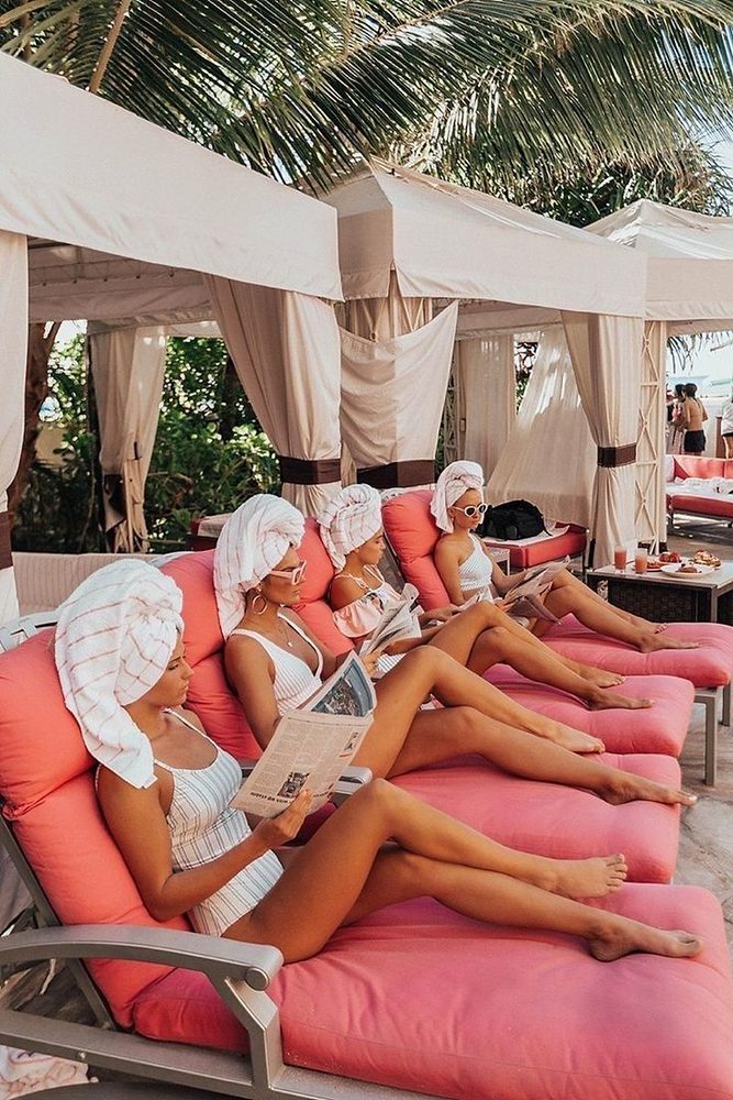Four friends in towel turbans reading magazines on pink loungers under white cabanas