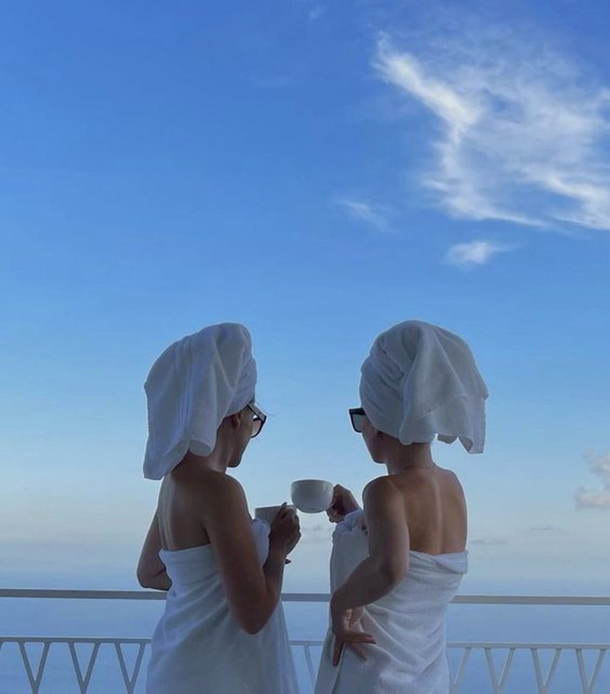 Two friends in towels clinking coffee cups on a balcony high above a calm blue sea
