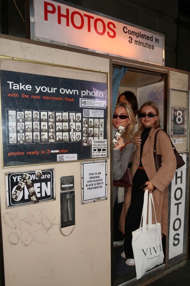 Three friends in coats squeezed into a vintage street photobooth at night, one holding up a photo strip