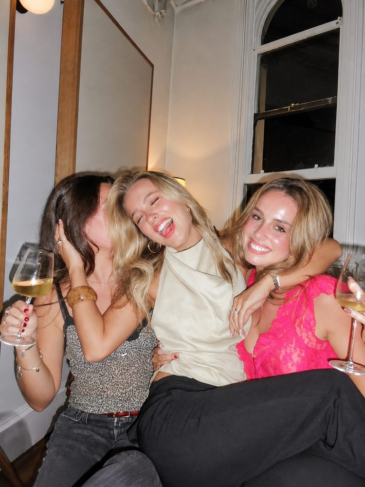 Three friends squeezed together laughing with glasses of white wine on a night out
