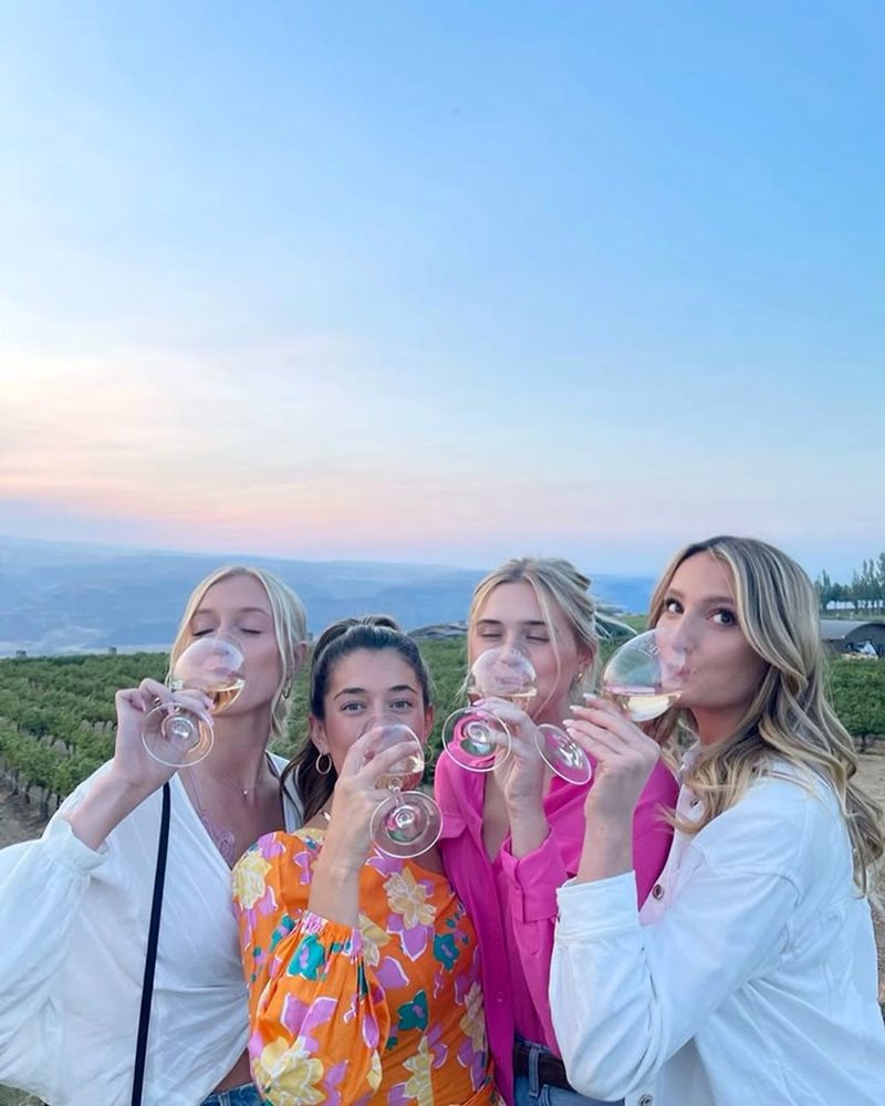 Four friends sipping rose in unison with vineyard rows and hills behind at dusk