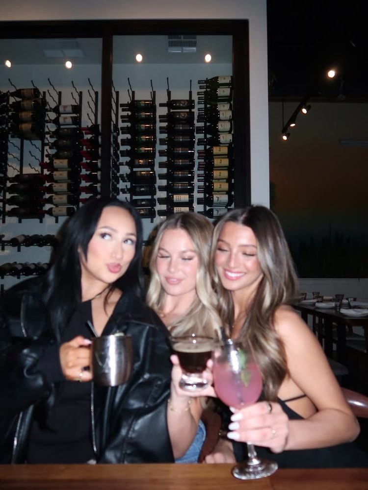 Three friends holding cocktails and a beer in front of a wine wall
